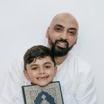 A father and son smiling, holding a holy book while wearing traditional attire.